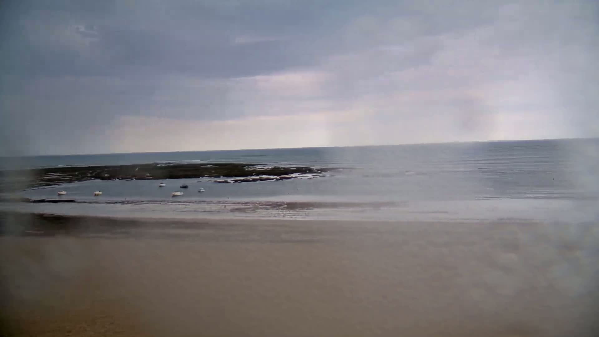 A wide, sandy beach stretches to a calm sea under an overcast sky, with several small boats visible in the shallows.