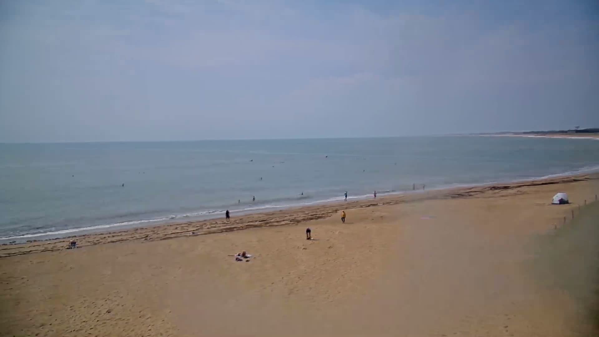 A mostly sunny day at a beach shows a calm ocean with a few people swimming and several people relaxing on a wide sandy beach.