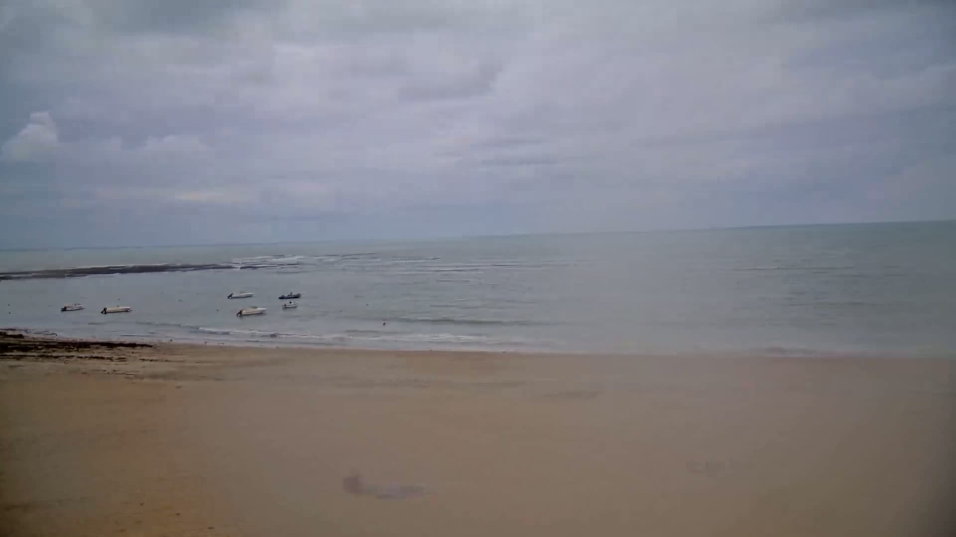 A sandy beach stretches to a calm, gray sea under an overcast sky, with several small boats anchored in the shallow water.
