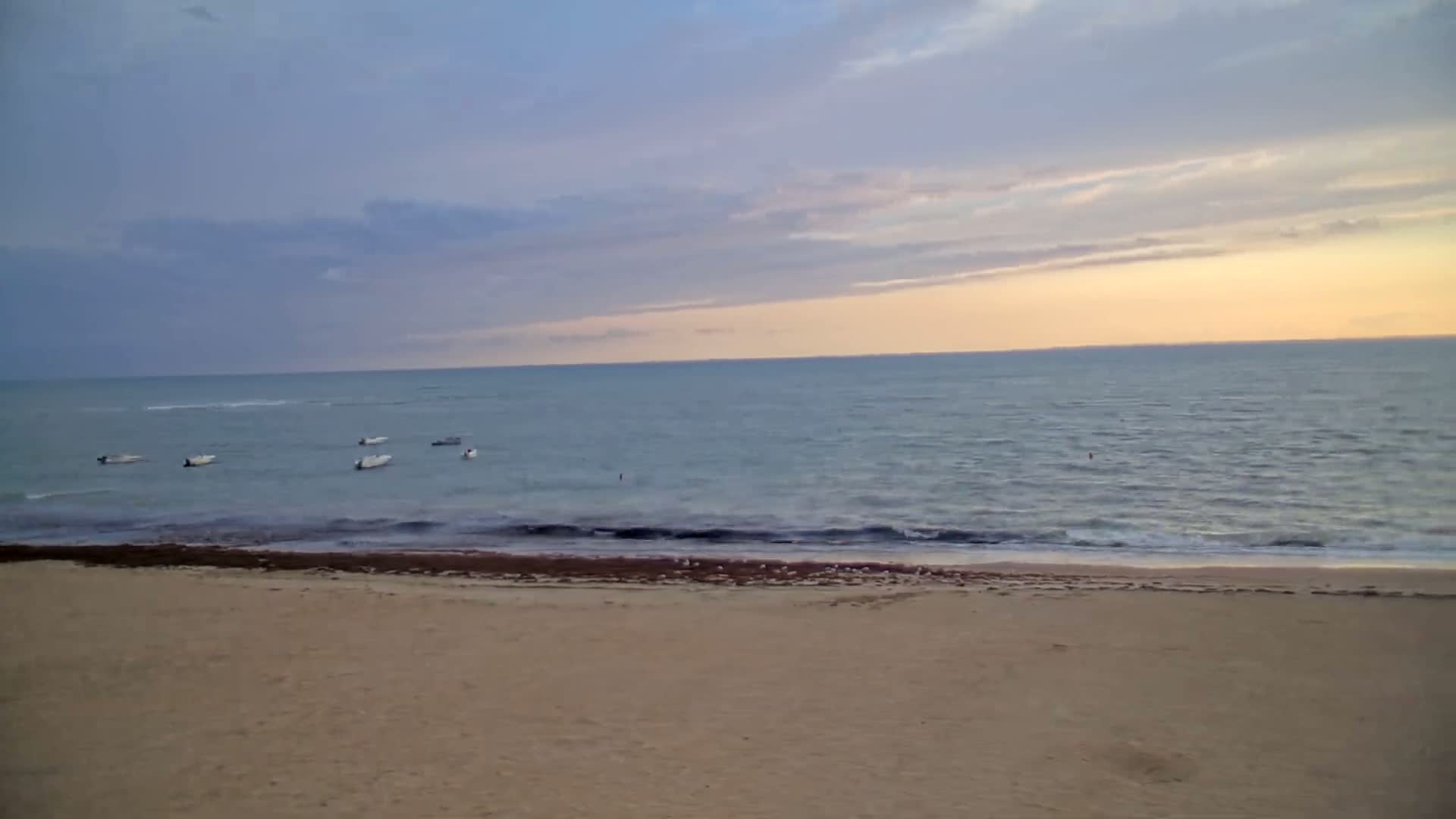 A sandy beach stretches to a calm, gray sea under an overcast sky, with several small boats anchored in the shallow water.