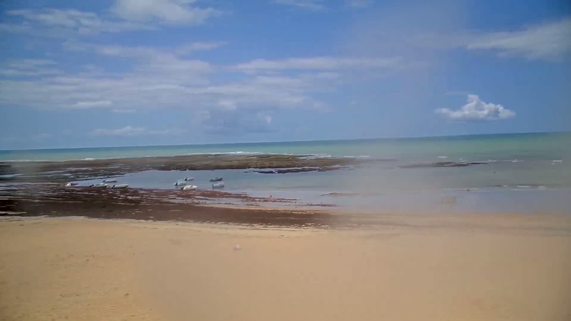 A sandy beach stretches out to a calm ocean where several small boats are anchored near a rocky outcrop under a mostly sunny sky.