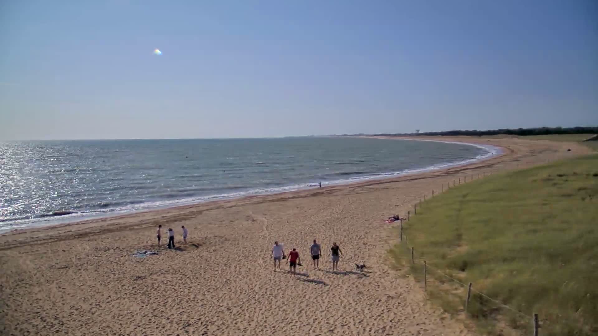 A sandy beach curves along a calm, blue ocean under a clear, sunny sky, with several people walking and relaxing on the shore.