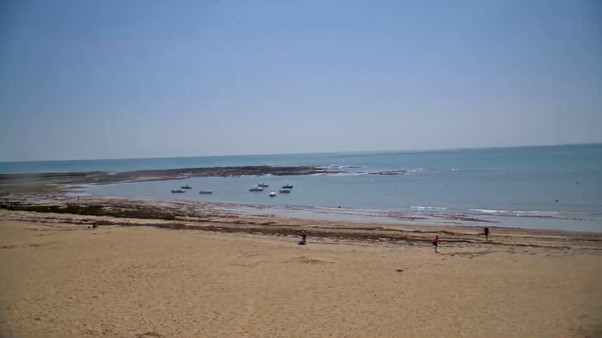 A sandy beach slopes down to calm, light blue water where several small boats are anchored near a rocky outcropping, under a clear, sunny sky.