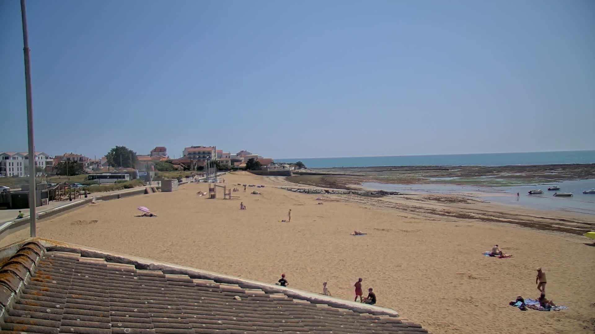 A sandy beach with a few people sunbathing and swimming, a town in the background, and calm ocean waters under a clear blue sky.