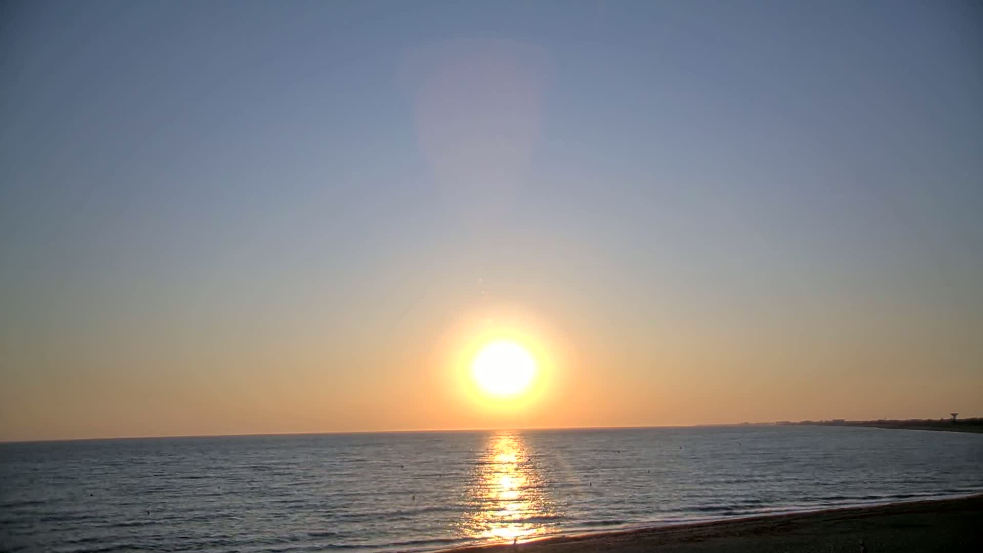A large, bright sun sets over a calm ocean under a clear sky.