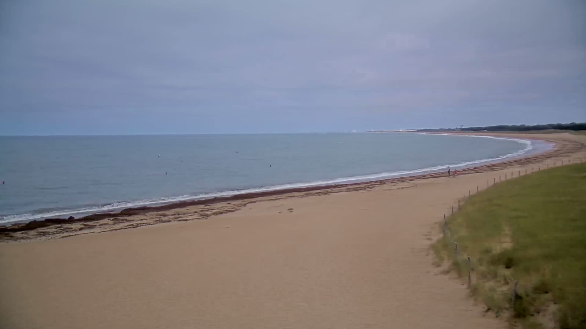 A sandy beach curves gently around a calm, gray ocean under an overcast sky.