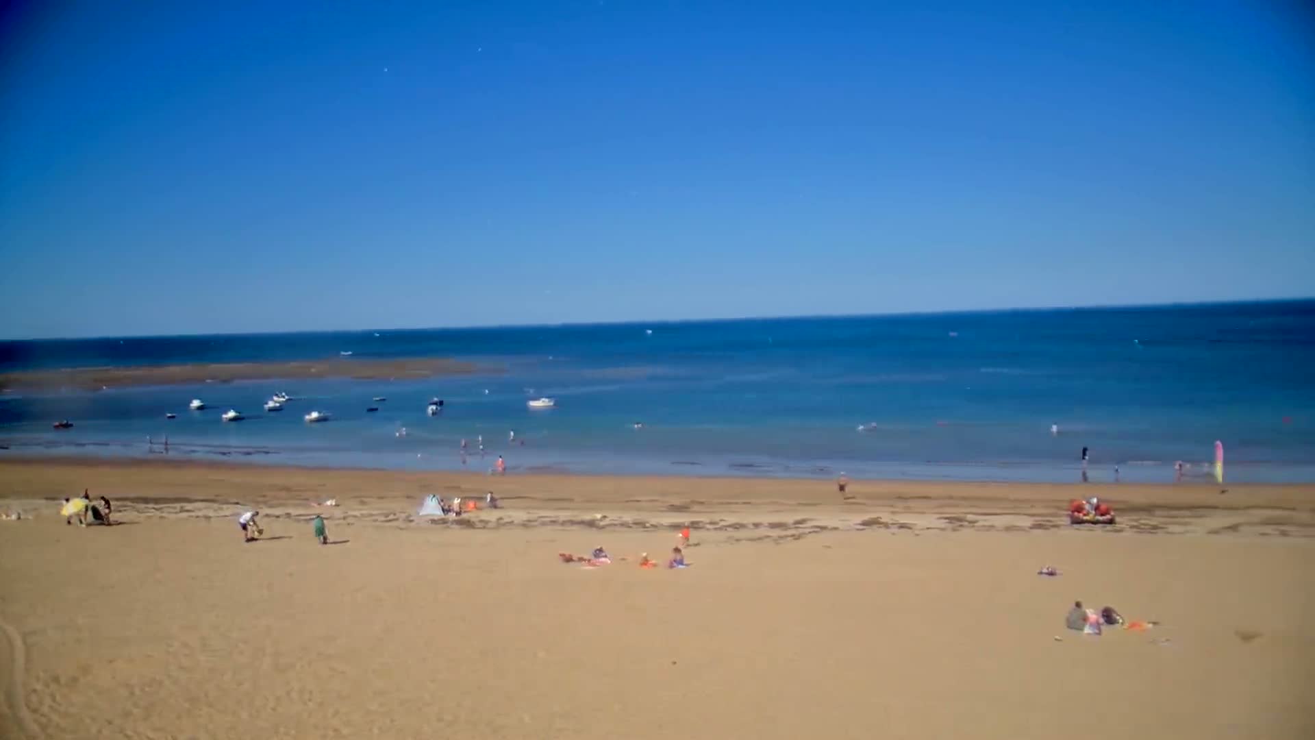 A sandy beach with scattered people and several small boats in calm, clear blue water under a bright blue sky.