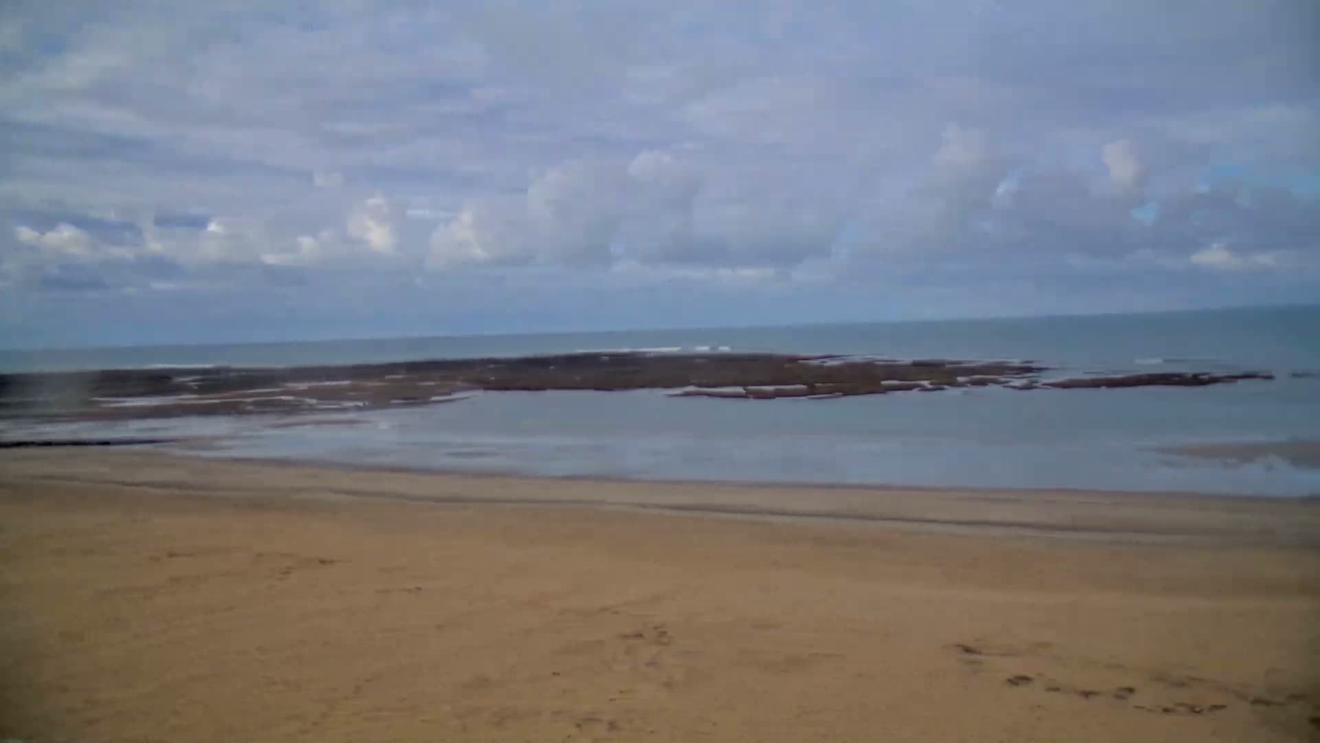 A sandy beach extends to a calm sea dotted with rocky formations under a cloudy sky.