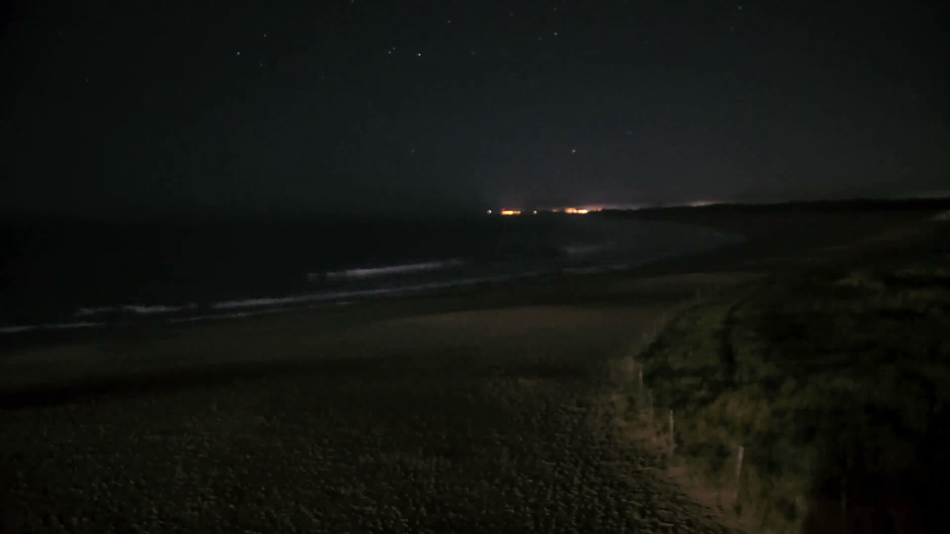 A sandy beach extends to a calm sea dotted with rocky formations under a cloudy sky.
