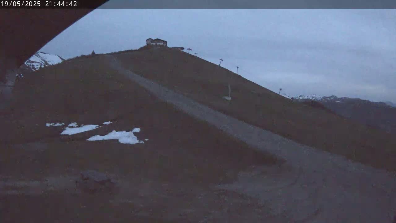 A dirt road winds down a dark, snow-dusted mountainside under a gloomy sky, passing a small building and ski lift towers.