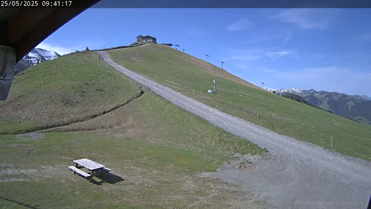 A gravel road winds up a grassy hillside towards a small building under a clear blue sky, with snow-capped mountains visible in the distance.