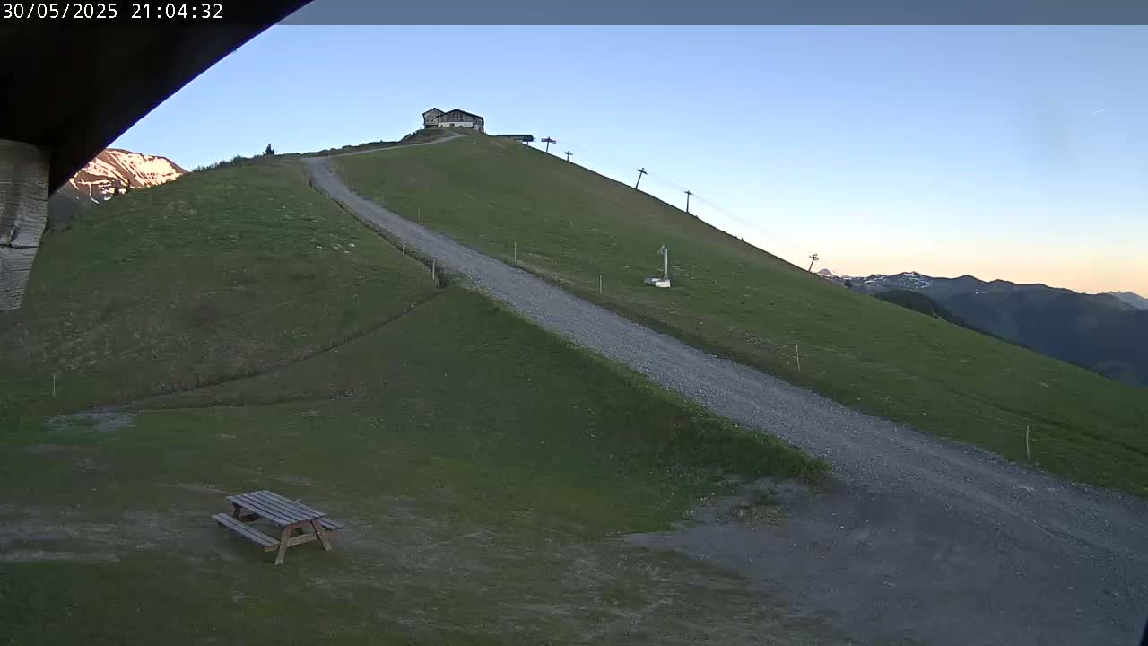 A gravel road winds down a grassy hillside toward a picnic table, with a building and ski lift visible uphill under a clear twilight sky.