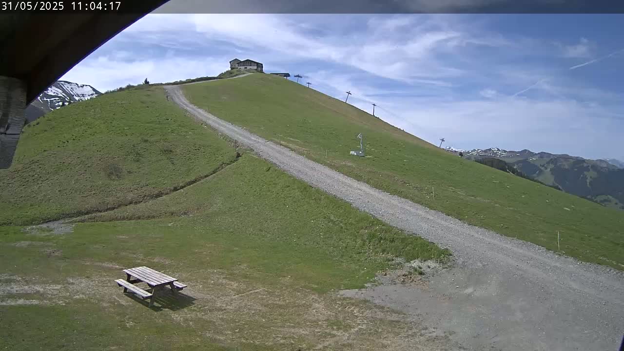 A gravel road winds down a grassy hillside toward a picnic table, under a partly cloudy blue sky, with a building and ski lift visible further up the hill.