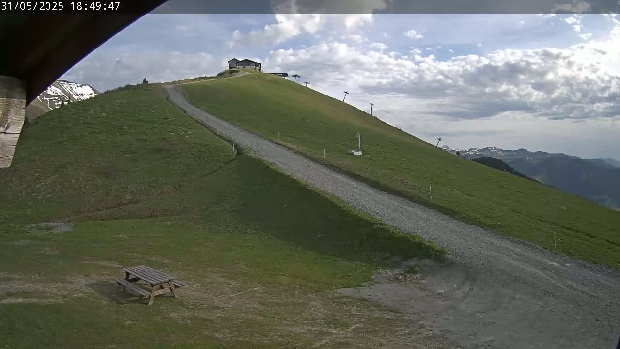 A gravel road winds down a grassy hill toward a picnic table, with a small building and ski lift visible higher up, under a partly cloudy sky.