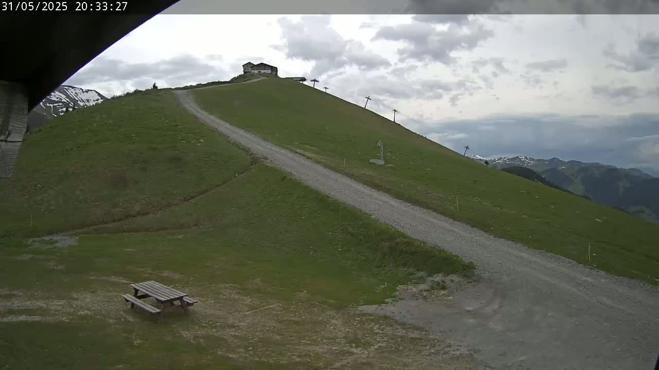A gravel road winds down a grassy hillside toward a picnic table, with a small building and ski lift visible higher up, under a cloudy sky.