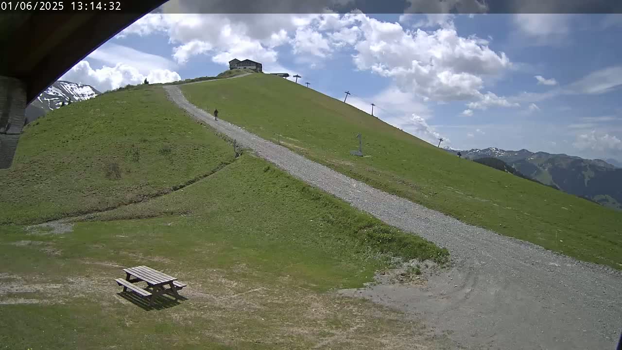 A gravel path winds down a grassy hillside towards a picnic table, under a partly cloudy sky, with a small building and ski lift visible at the top of the hill.
