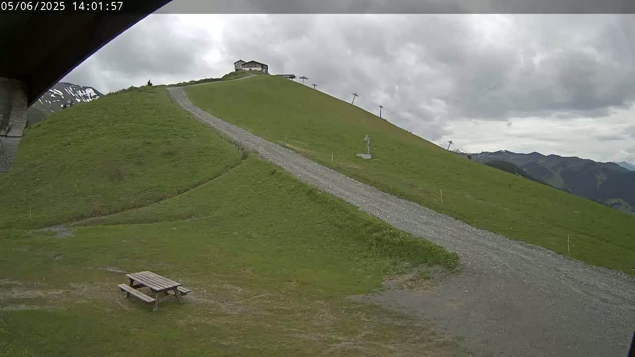 A gravel road winds down a grassy hill towards a picnic table, with a building and ski lift visible further up the hill under an overcast sky.