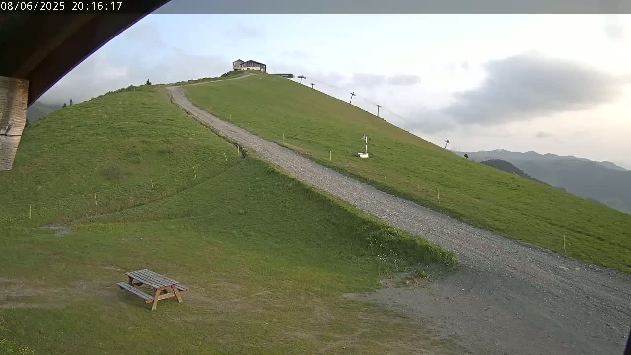 A gravel road winds down a grassy hill towards a picnic table, with a building and ski lift visible further up the hill under a cloudy sky.