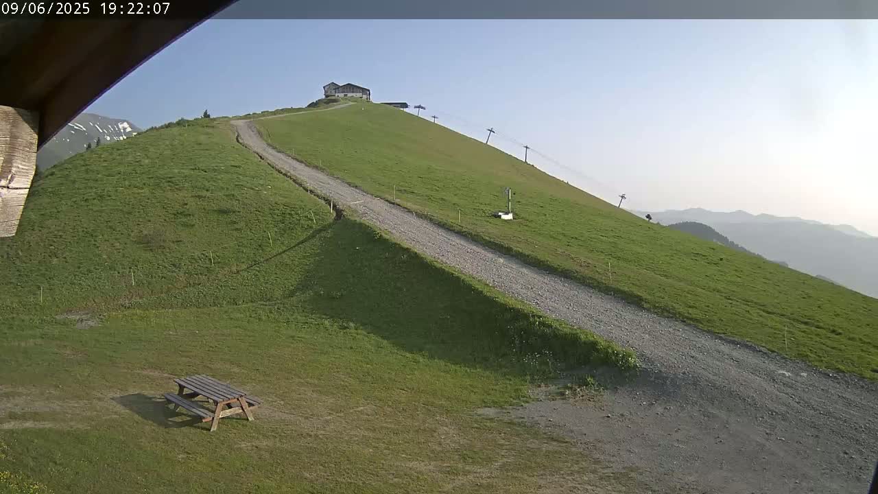 A gravel road winds up a grassy hill toward a small building under a clear sky, with a picnic table situated at the base of the hill.