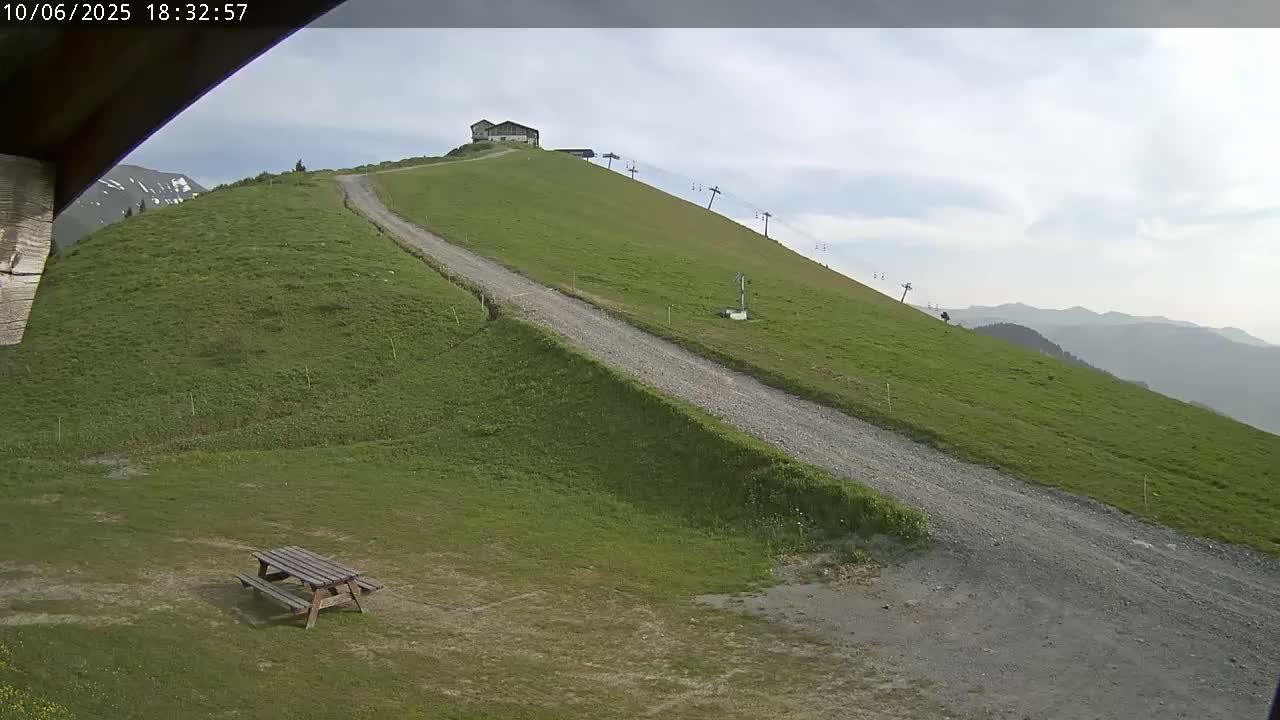 A gravel road winds up a grassy hillside towards a small building and a chairlift, under a mostly cloudy sky.