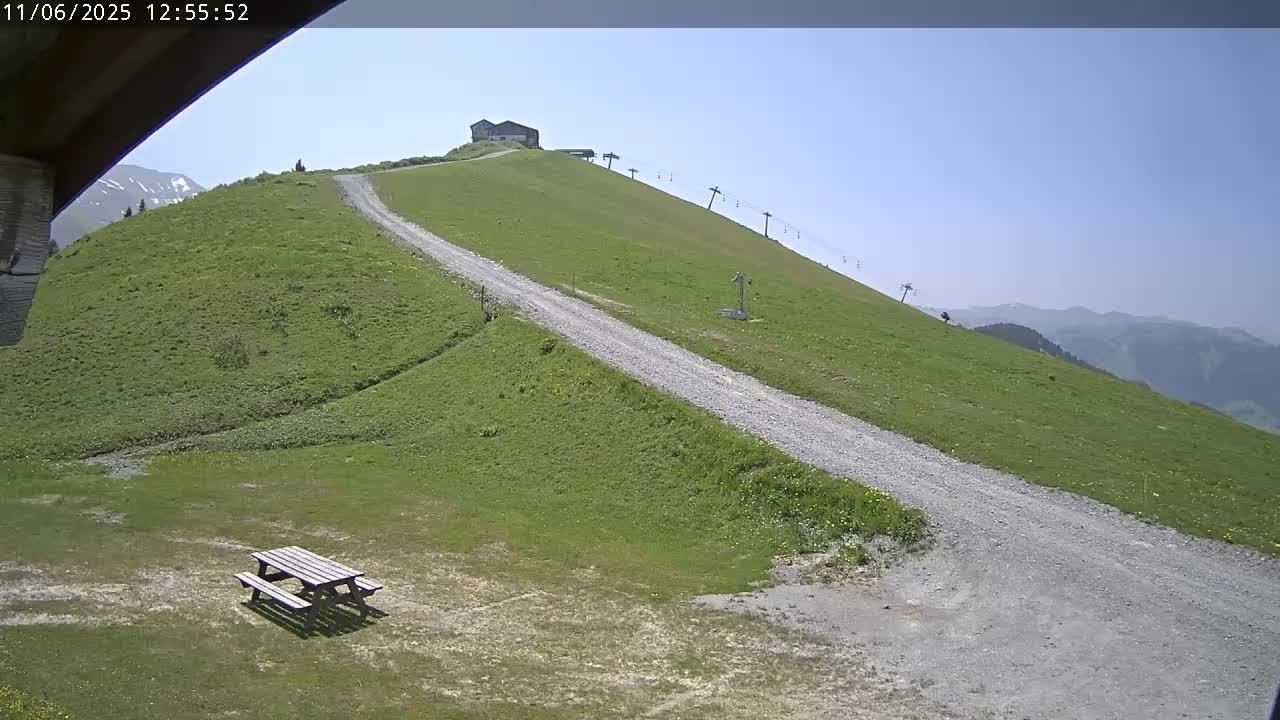 A gravel road winds down a grassy hill towards a picnic table, with a small building and chairlift visible further up the hill under a clear blue sky.