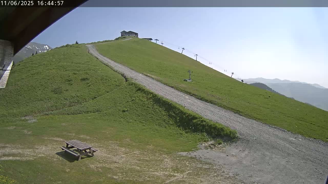 A grassy hillside with a gravel road leading up to a small building, a chairlift, and a picnic table in the foreground, under a clear sunny sky.