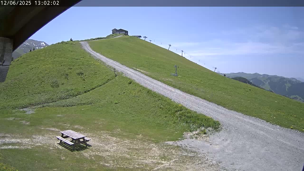 A gravel road winds down a grassy hill toward a picnic table, with a chairlift and a small building visible further up the hill under a clear blue sky.