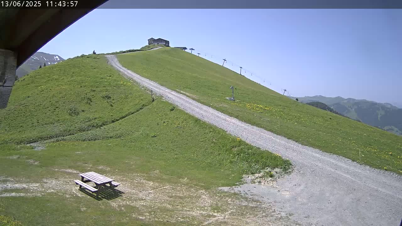 A gravel road winds up a grassy hillside on a clear, sunny day, passing a picnic table and a small building near the top, with a chairlift visible in the background.