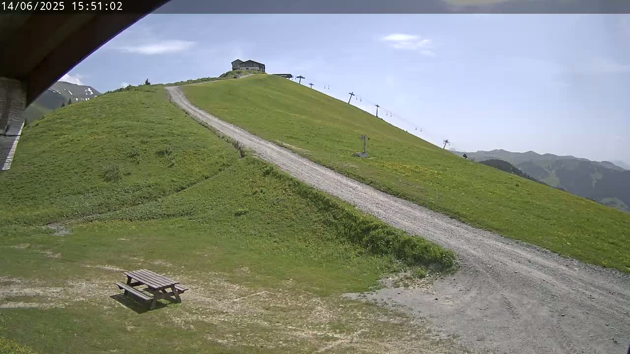 A grassy hillside with a gravel road leading up to a small building, a chairlift, and a picnic table in the foreground under a mostly sunny sky.
