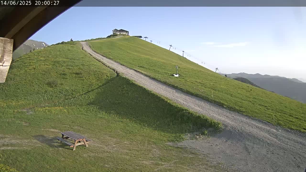 A grassy hillside with a gravel road leading up to a small building, a chairlift, and a picnic table in the foreground under a mostly sunny sky.