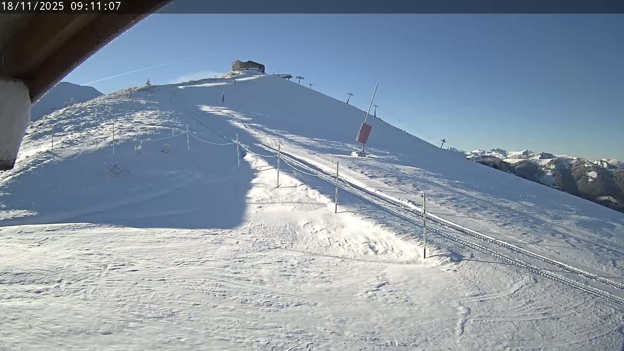 A clear and sunny winter day illuminates a snow-covered mountain ski slope with a building at the peak, ski lifts, and distant snowy mountain ranges.