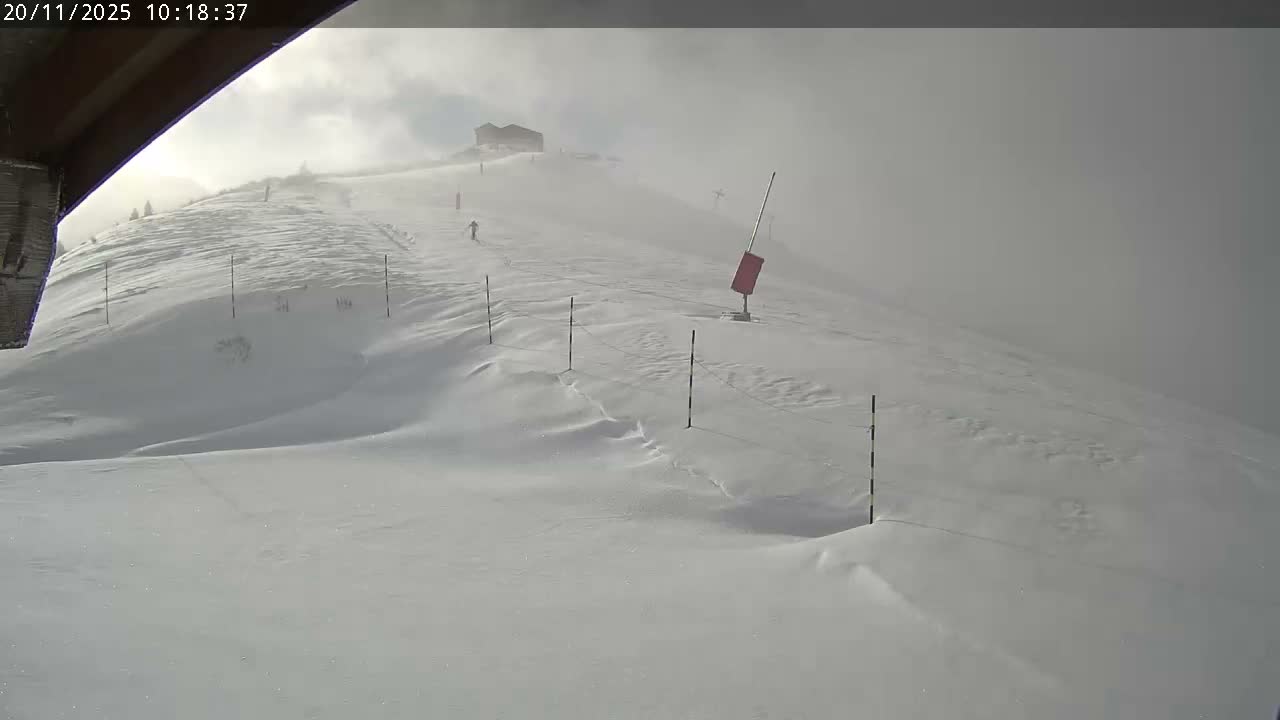 A snow-covered mountain slope is enveloped in heavy fog and overcast skies, with a distant building and marker poles barely visible amidst the blowing snow.