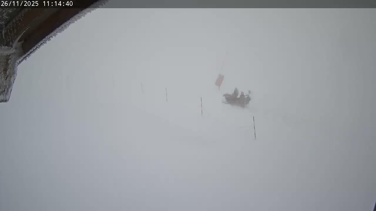 A snowy outdoor scene displays a frost-covered building edge in the upper left, while a snow vehicle with a few people and a red marker is faintly visible in the distance, all under severe whiteout conditions.