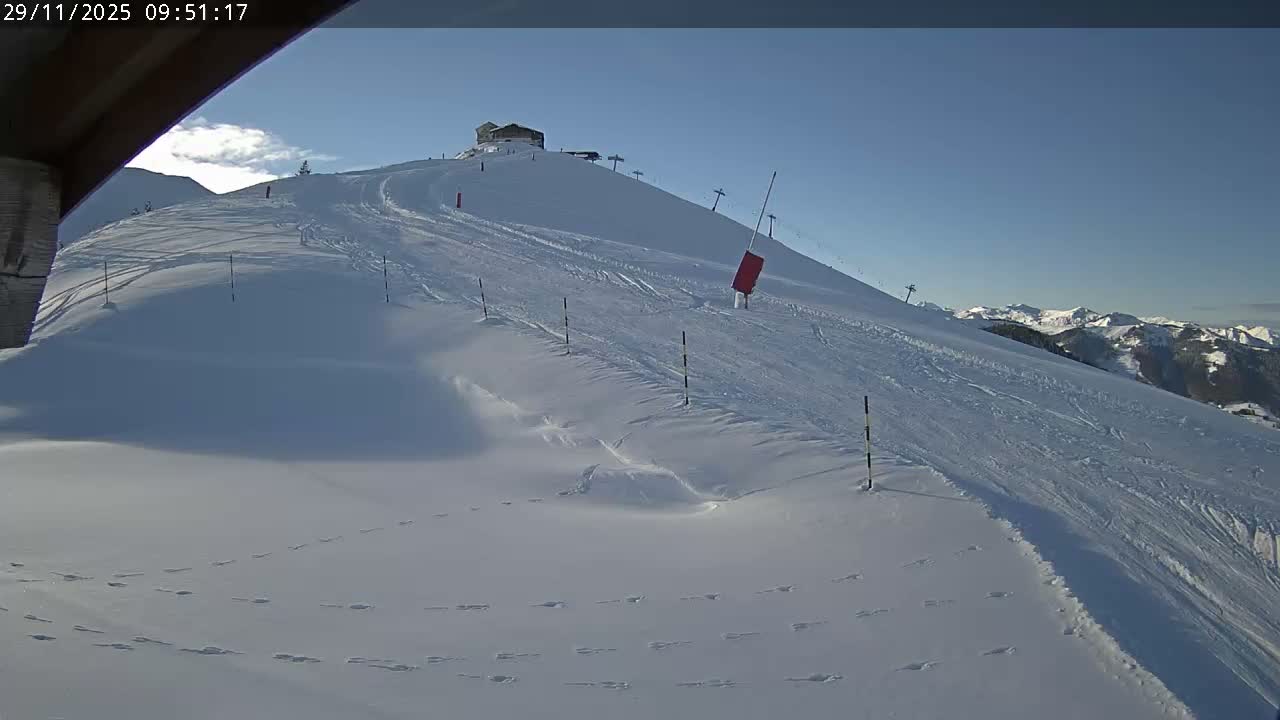 A snow-covered mountain ski slope with a building at its peak and several ski lifts is captured under a clear blue sky, showcasing ski tracks and footprints in the fresh snow, with distant snow-capped mountains visible in the background.