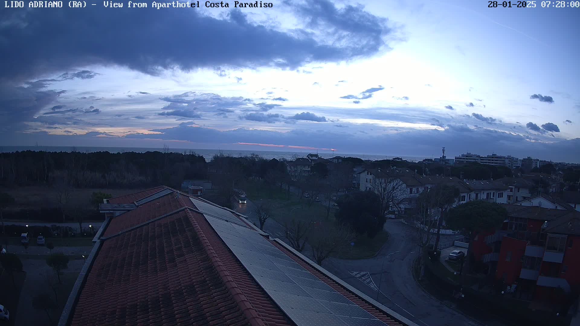 A mostly cloudy, dusky sky over a coastal town with a view of the sea from a rooftop with solar panels.