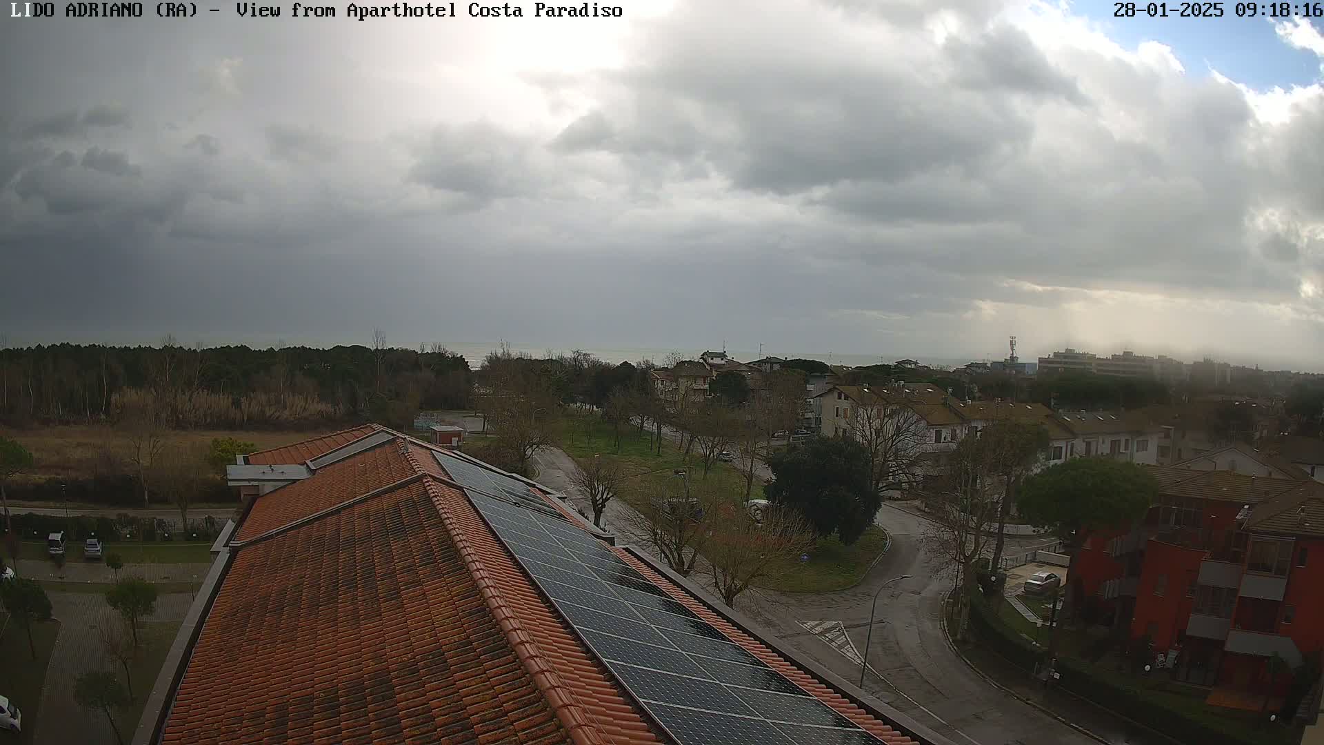 An overcast sky looms over a residential area with a coastal view, partially obscured by trees, and a building with solar panels on its roof.