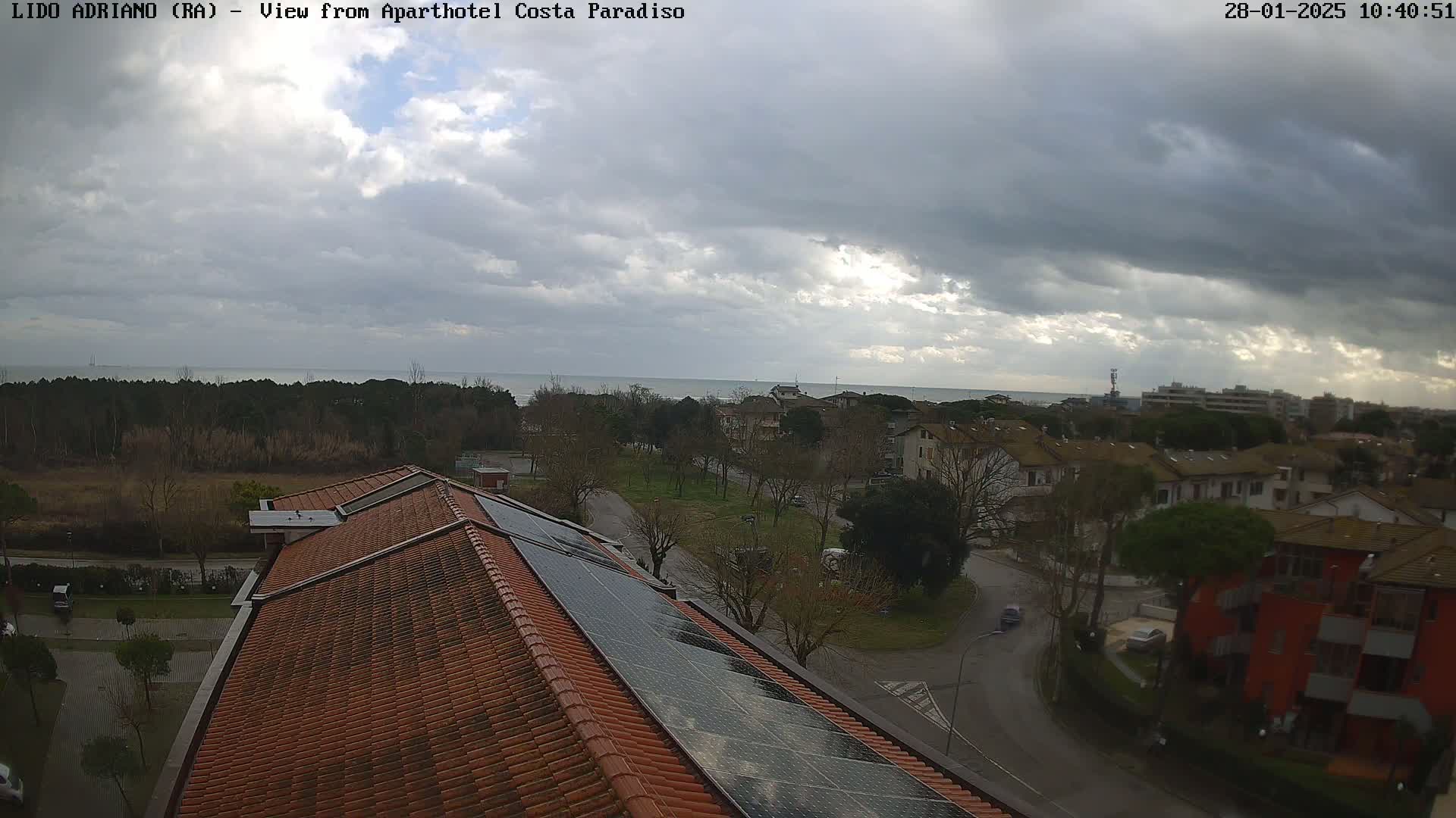 A mostly cloudy day overlooks a coastal town with a red building and solar panels on a brown roof in the foreground.