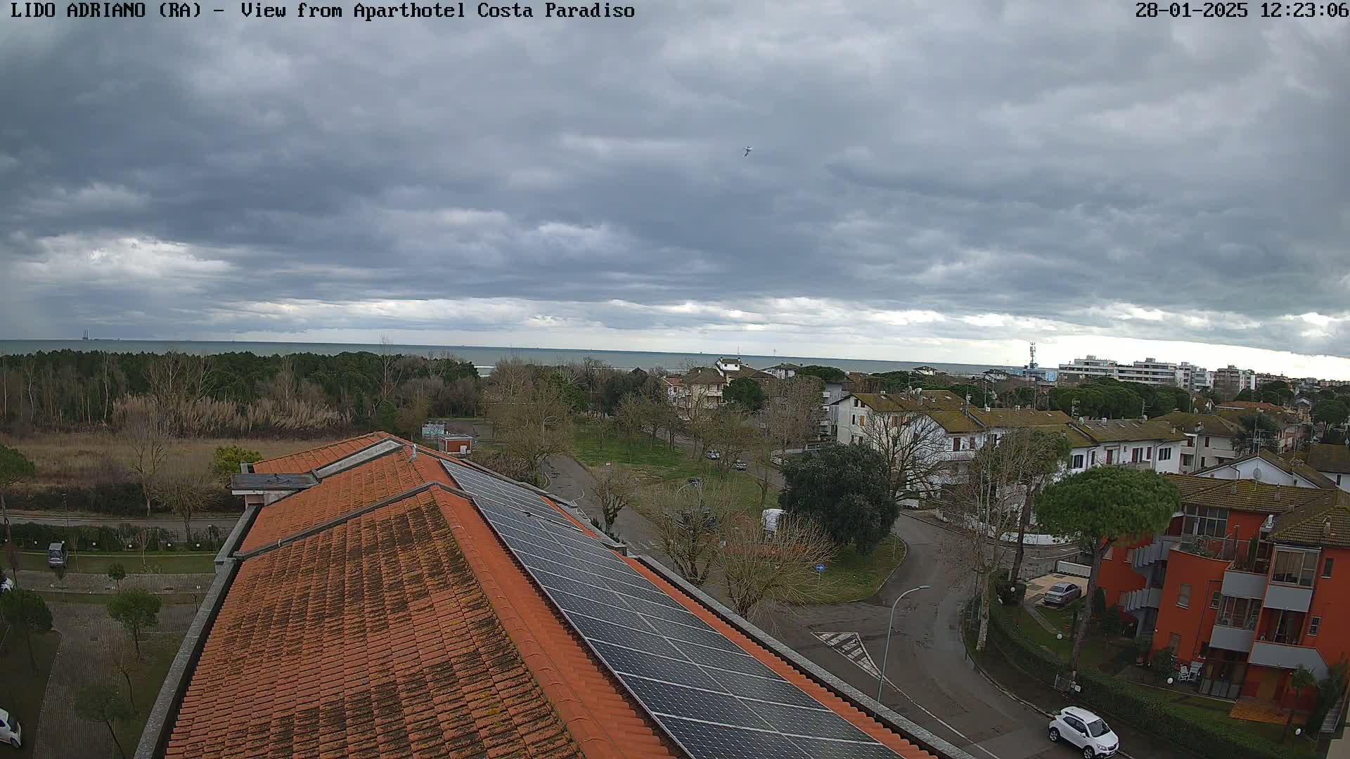 A view of an overcast day shows a coastal town with orange-tiled rooftops and solar panels, overlooking a calm sea and a line of trees.