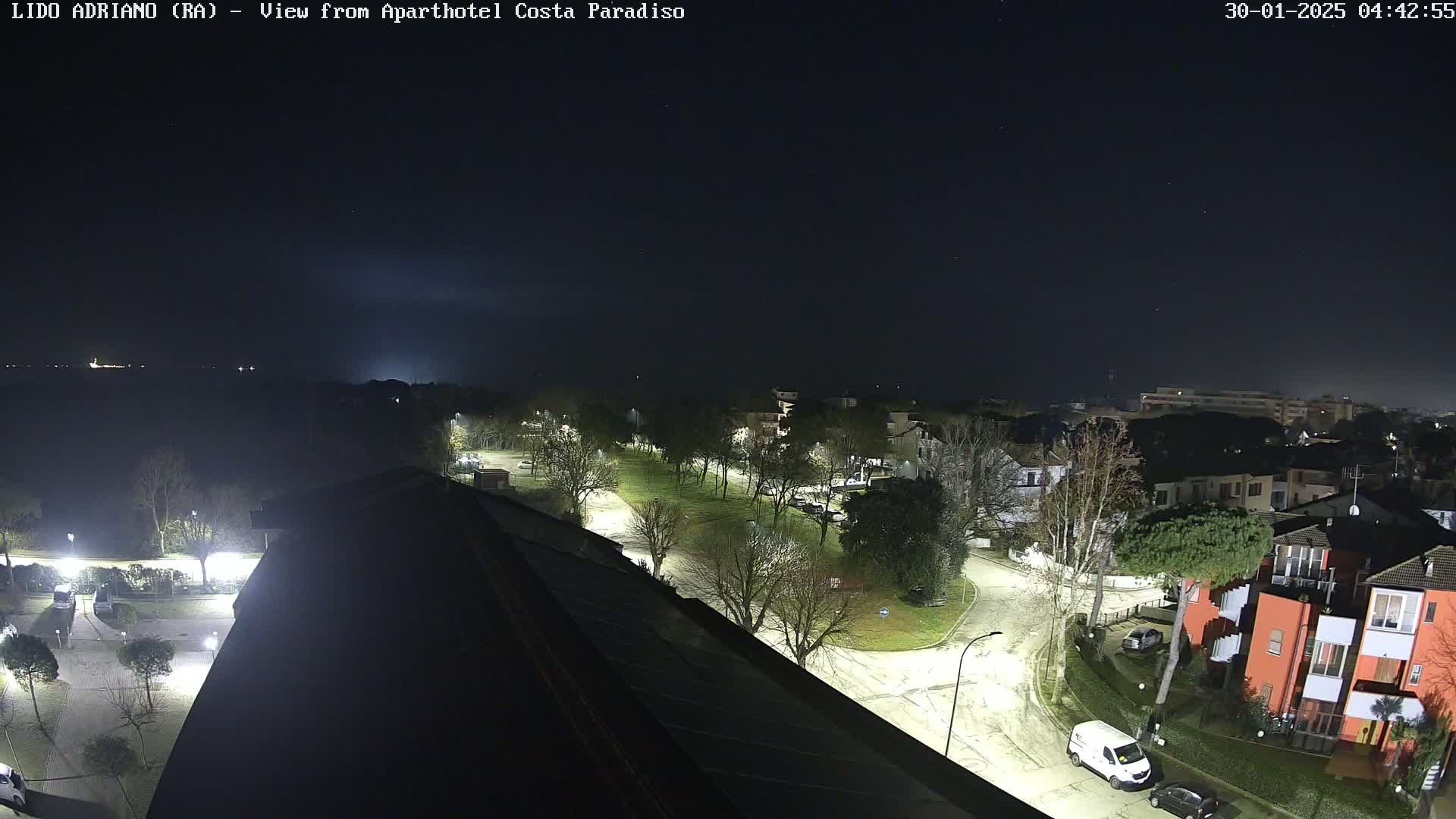 A nighttime aerial view of a residential area with a park, illuminated streets, and a few vehicles, under a clear, dark sky.