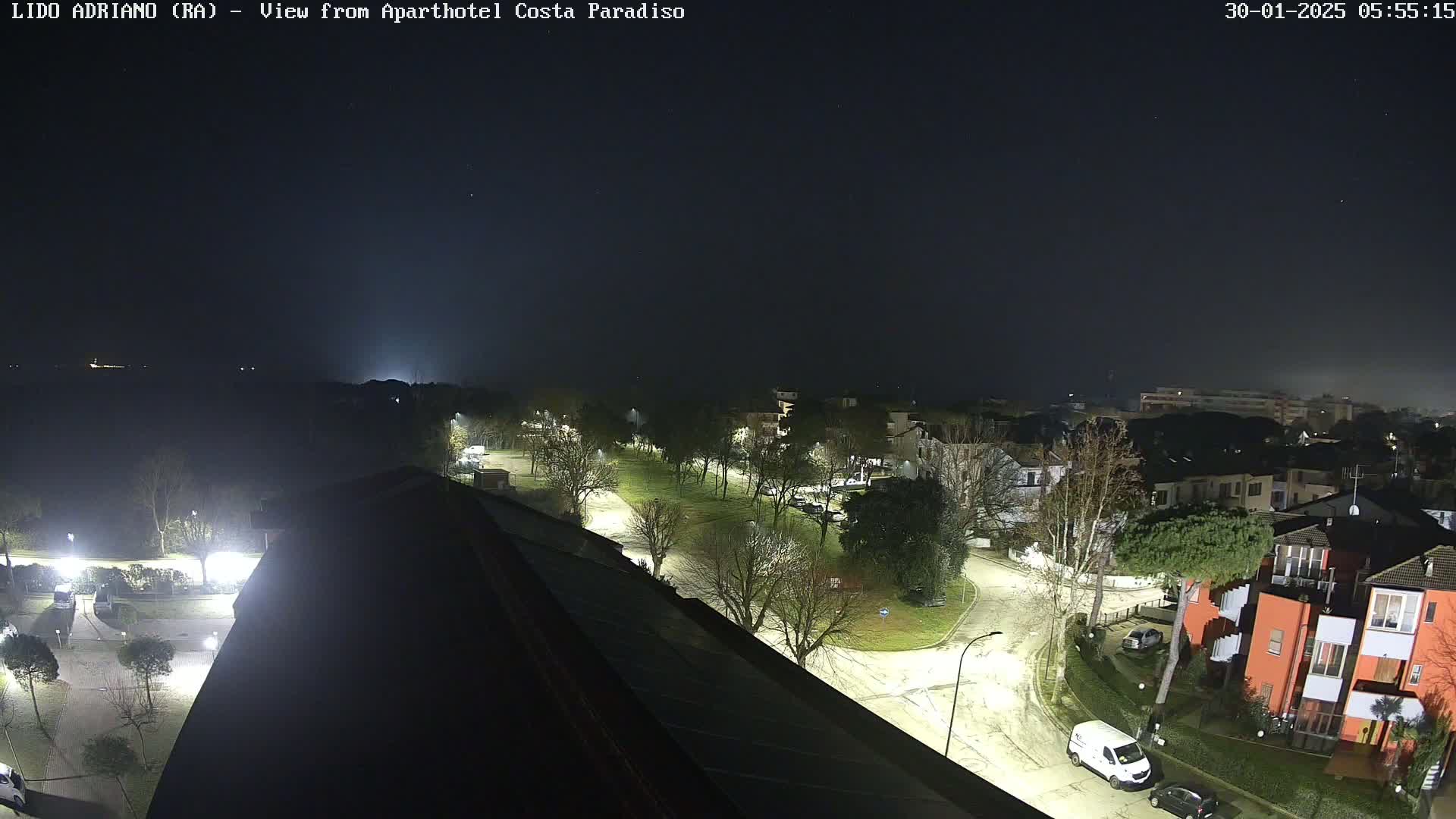 A nighttime aerial view of a residential area with streets and trees illuminated by streetlights under a dark, possibly foggy sky.