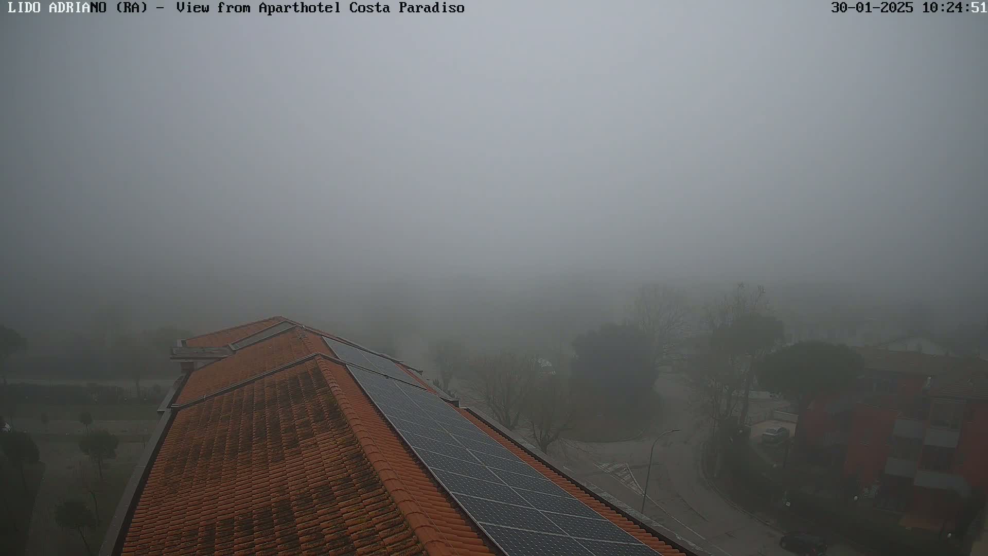 A thick fog obscures a view of a building's orange tiled roof with solar panels, overlooking a street and other buildings in the distance.