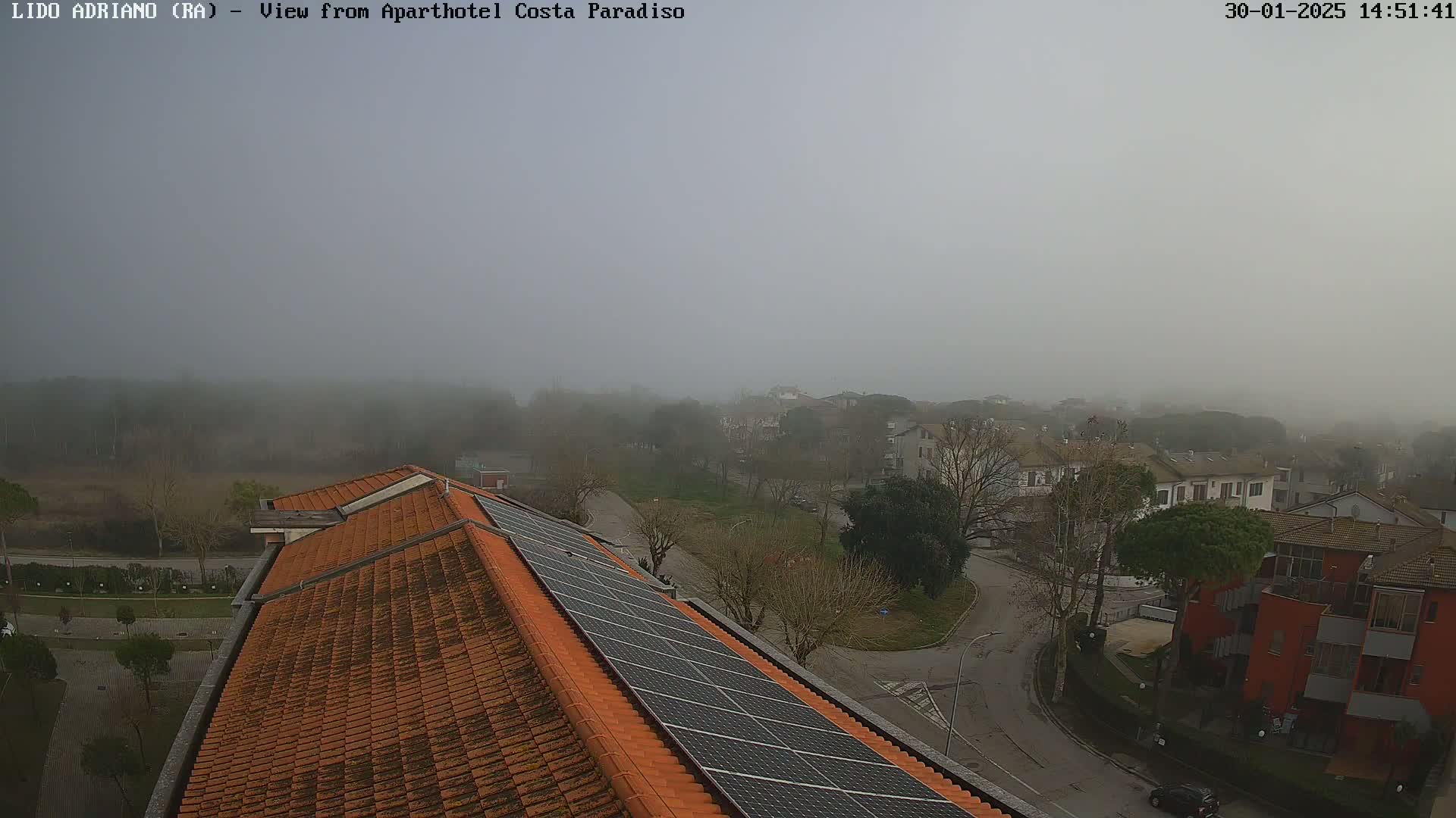 A foggy view from a rooftop with orange tiles and solar panels overlooks a residential area with roads and trees.