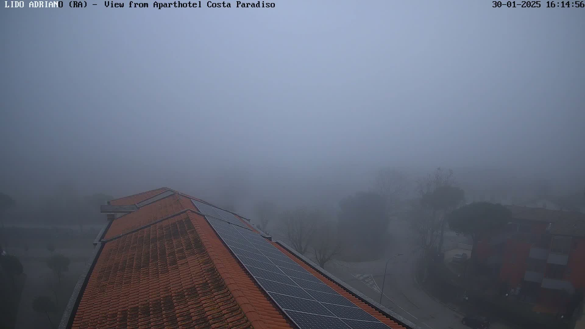A thick fog obscures a view of a building's red tile roof and solar panels, with hazy outlines of trees and buildings in the distance.