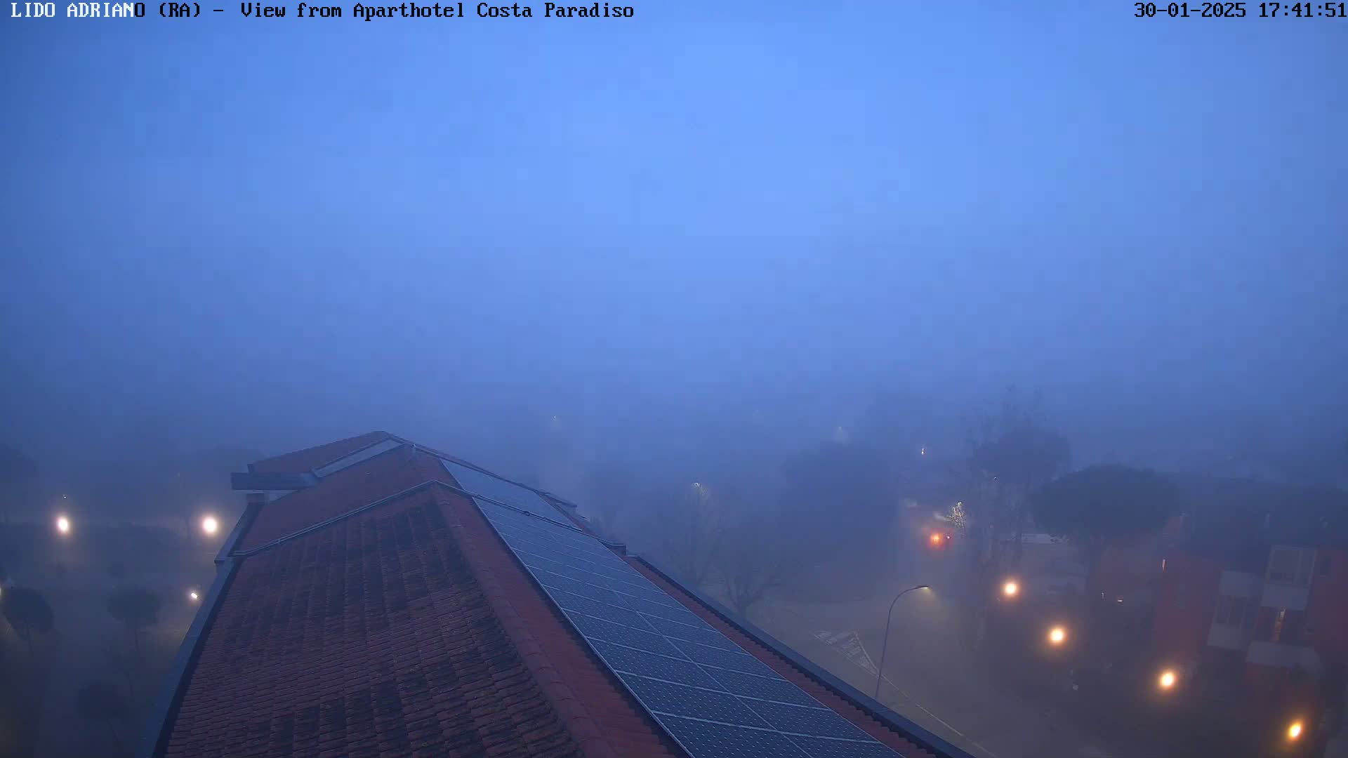 A rooftop with solar panels is visible under a thick, blue fog at dusk, with dimly lit buildings and streetlights obscured in the distance.