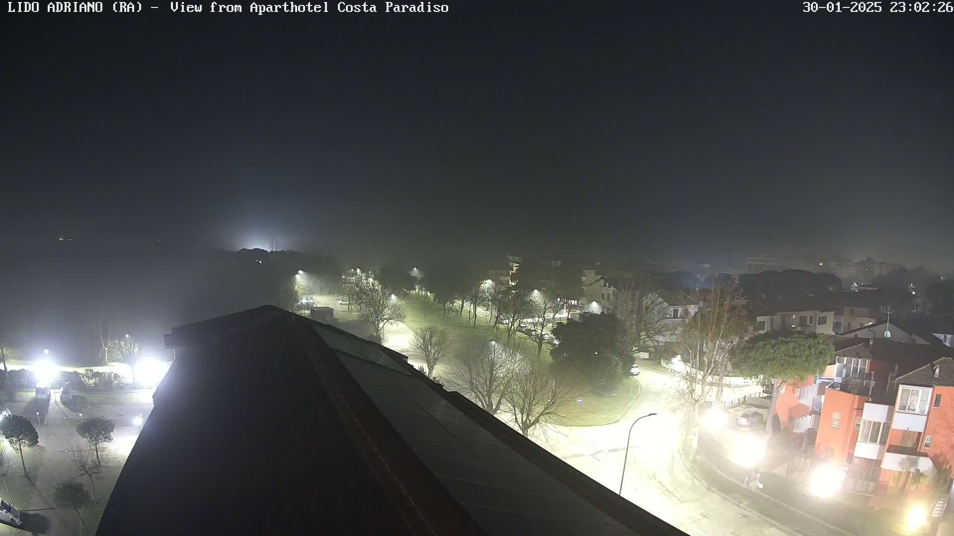 A foggy night scene shows a low-lit residential area with roads and trees, viewed from a rooftop.