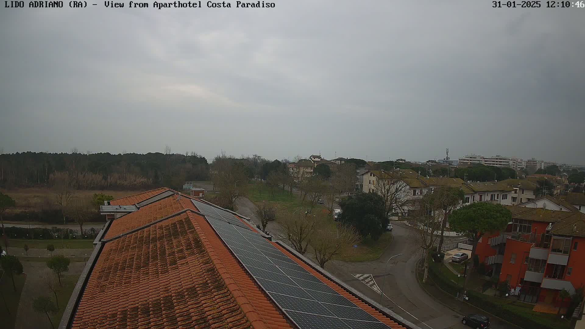 An overcast day reveals a view of a residential area with a red tile roof and solar panels, overlooking a street and other buildings from what appears to be a rooftop.