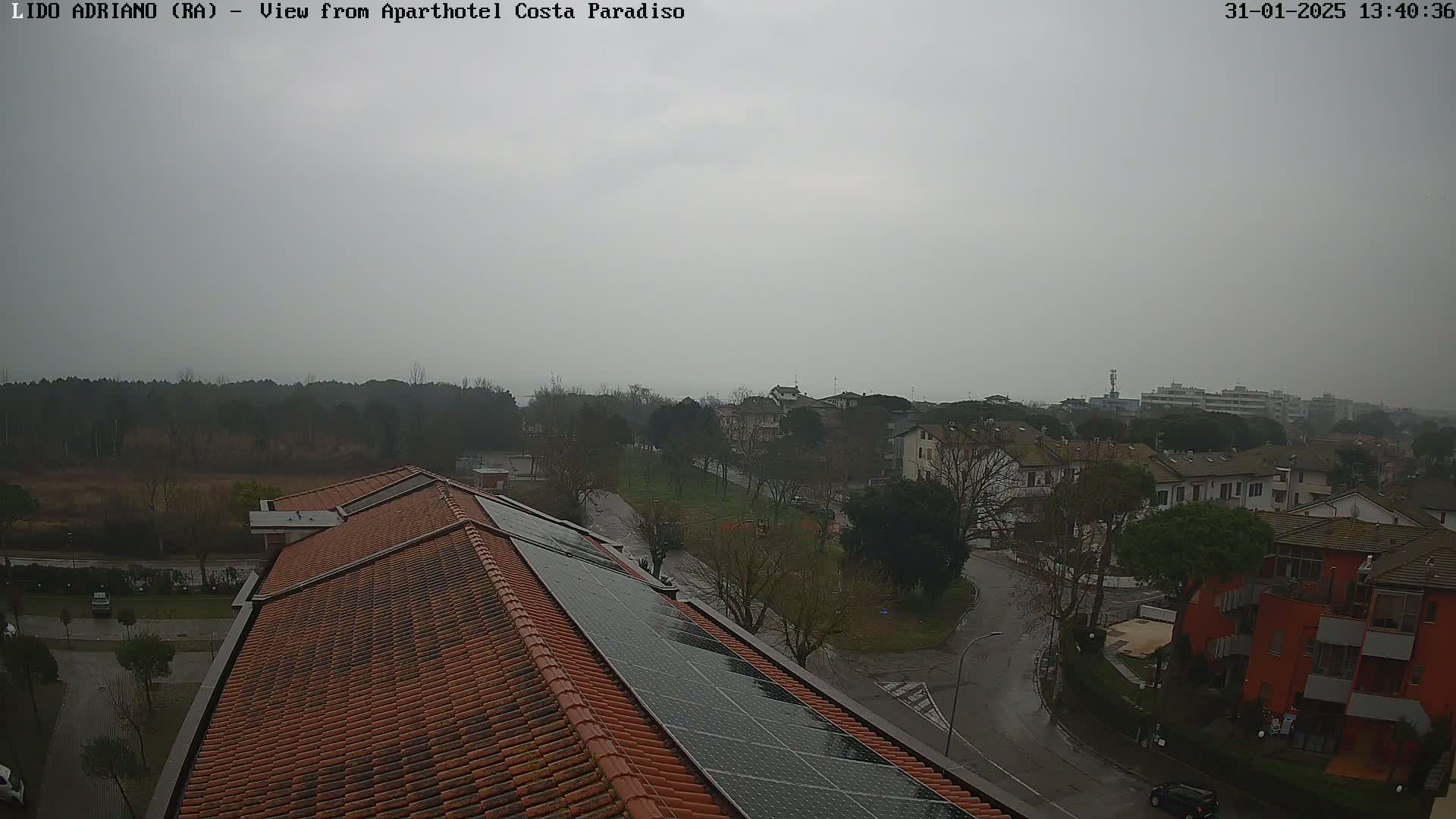 A high-angle view of a residential area on a cloudy, possibly rainy day, showing a building roof with solar panels and surrounding streets and houses.