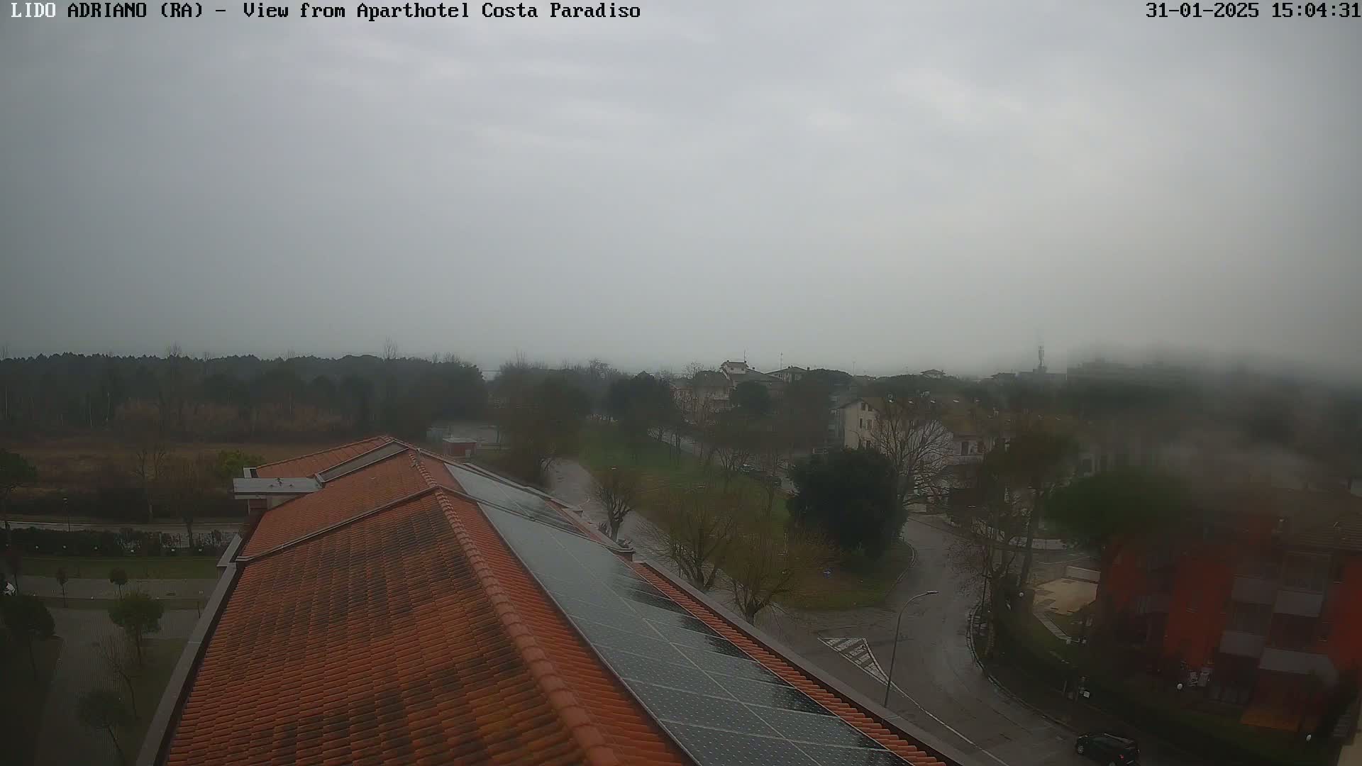 A view from a rooftop with orange tiles and solar panels shows a foggy, overcast town with trees and roads on a rainy day.