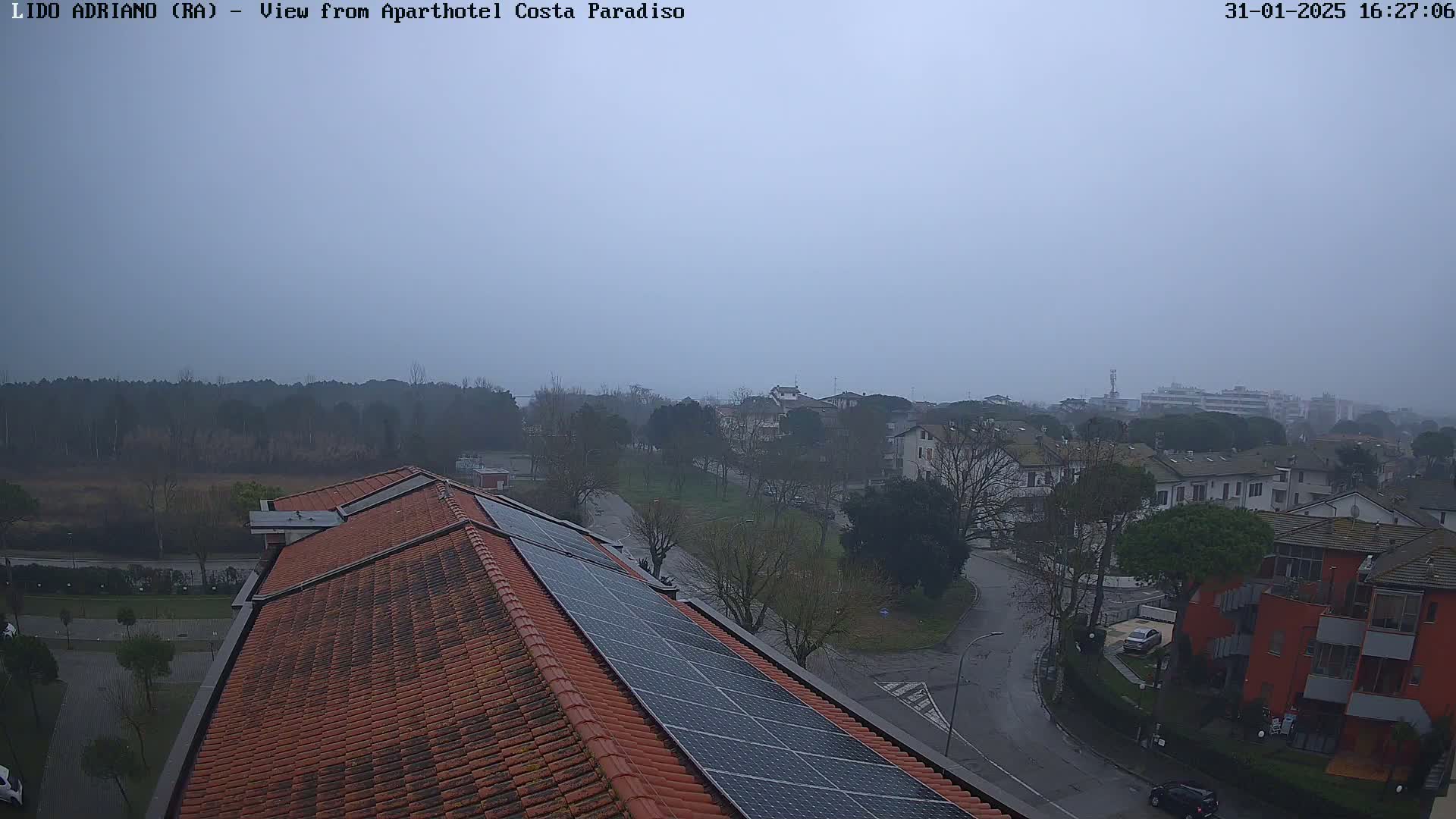 A hazy view from a rooftop with solar panels shows a residential area with low buildings and trees under an overcast sky.