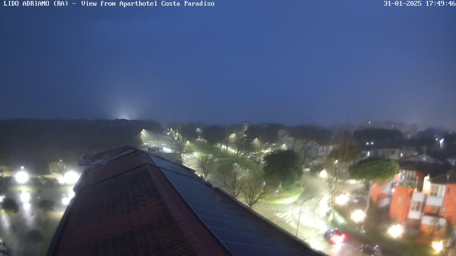 A nighttime view of a street scene shrouded in fog, showing a building with solar panels on its roof, illuminated streets, and nearby buildings.